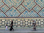 Two women and a child walk past a wall adorned with geometric tile patterns in Uzbekistan