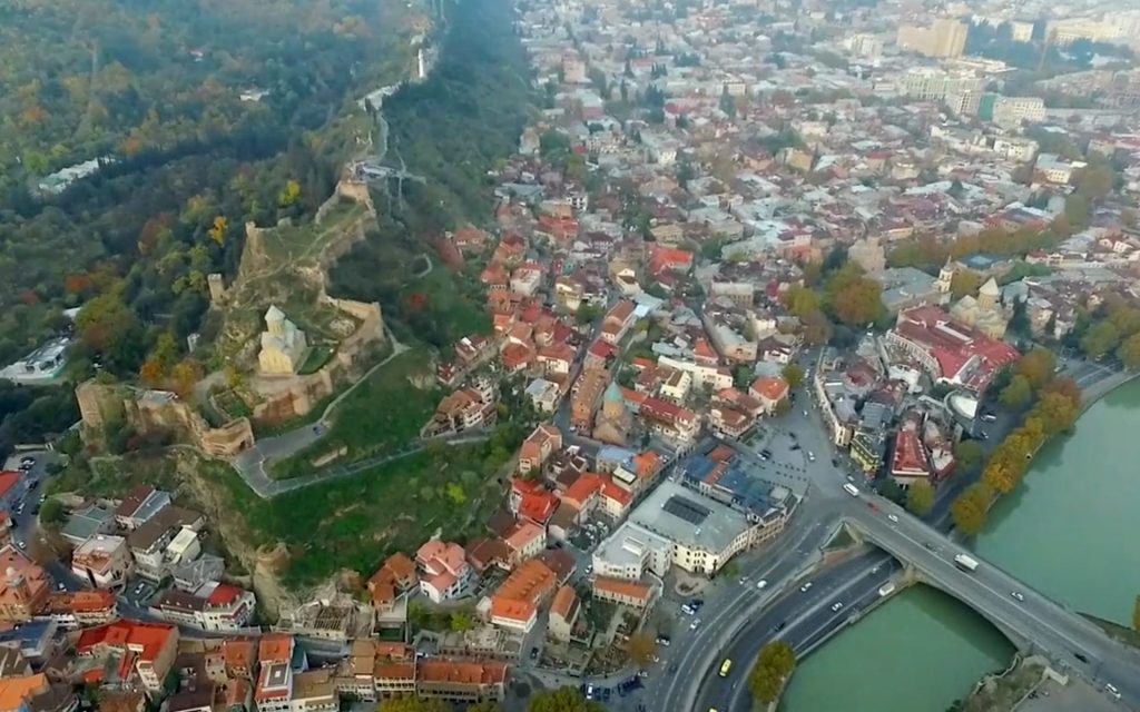 Aerial view of Tbilisi, showcasing the Narikala Fortress and a bridge crossing the Kura River amidst an urban landscape.