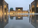 Registan square in Samarkand with ornate facades and domes is reflected symmetrically in a still water surface, illuminated at dusk. Photo credit: Abdu Samadov
