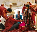 A group of people dressed in traditional Turkmen clothing gather around two seated individuals, engaging in a festive activity. Photo credit: Michel Behar.