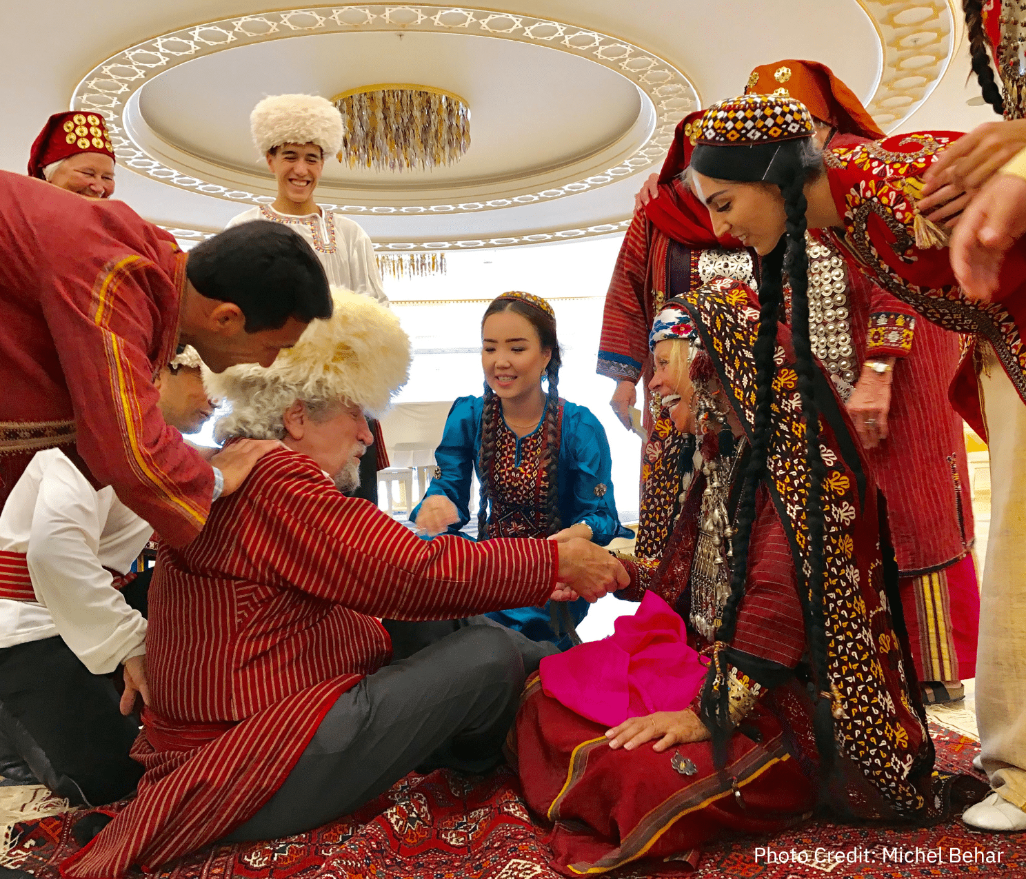 A group of people dressed in traditional Turkmen clothing gather around two seated individuals, engaging in a festive activity. Photo credit: Michel Behar.