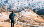 A person wearing a blue jacket and backpack takes a photo of a Gergeti Trinity Church in Kazbegi, Georgia