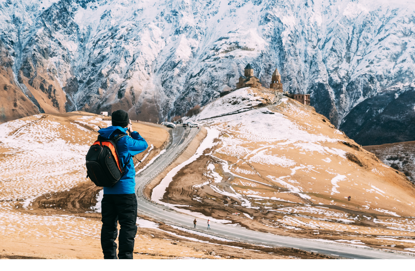 A person wearing a blue jacket and backpack takes a photo of a Gergeti Trinity Church in Kazbegi, Georgia